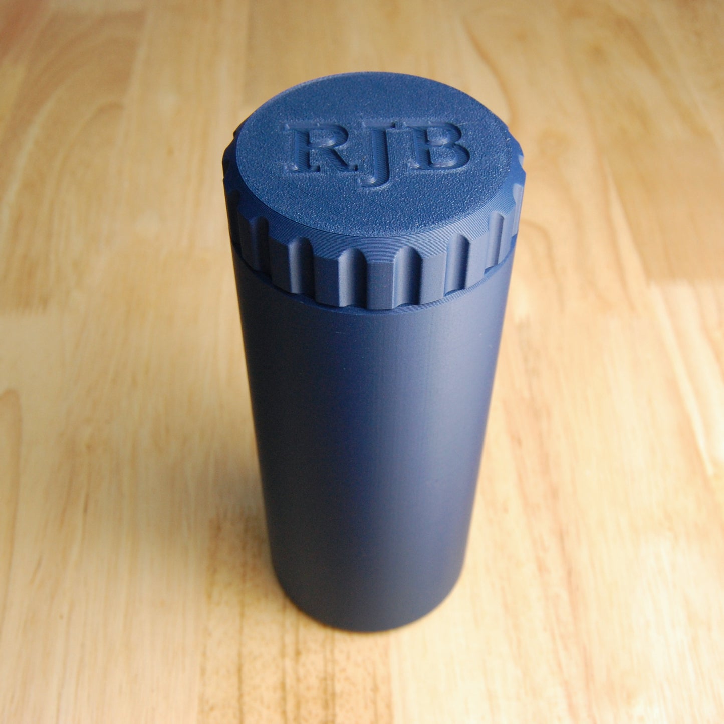 The capsule razor travel case in navy blue on a wooden background.  Personalized cap that says "RJB"