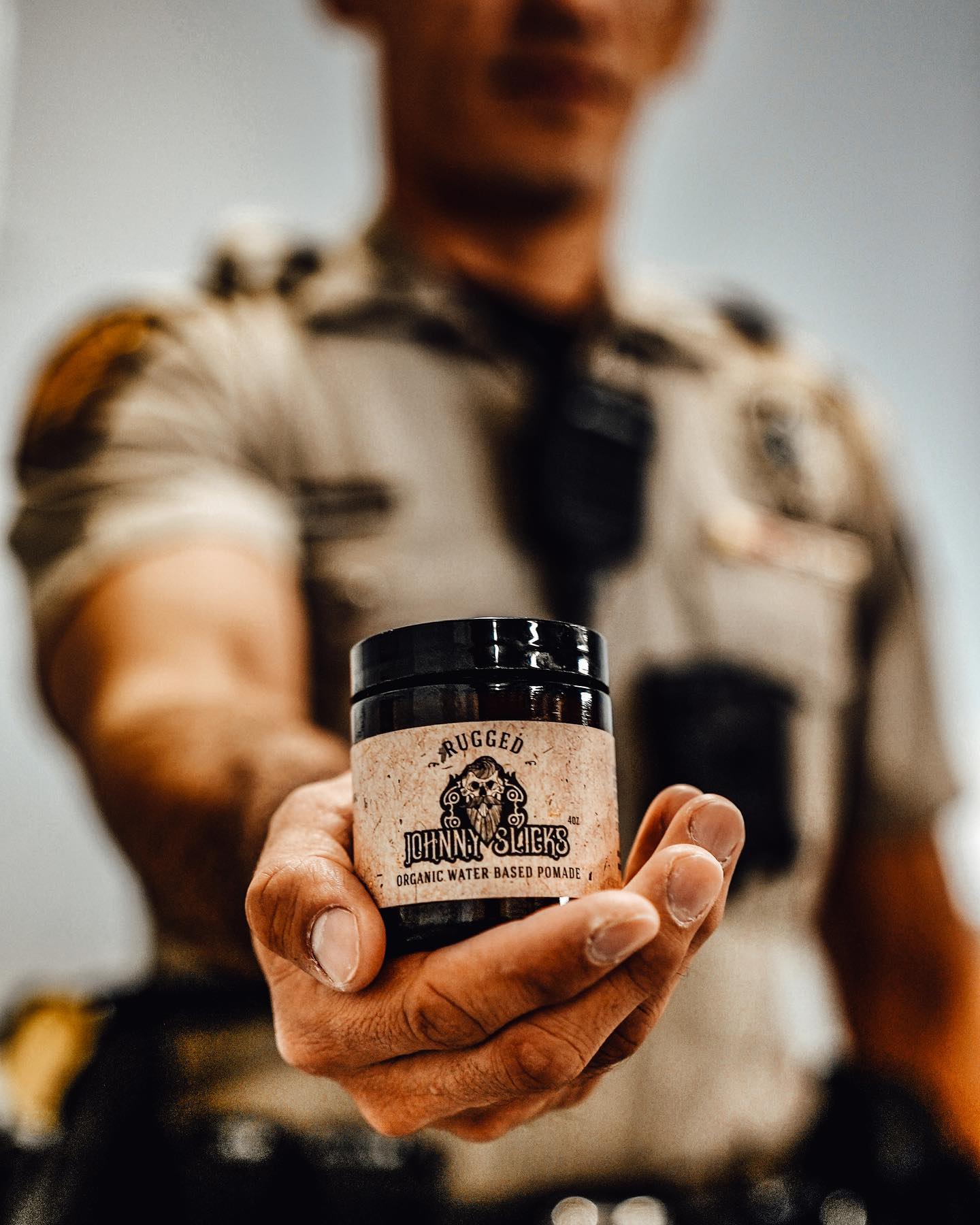 Man in uniform holding a jar of Johnny Slicks Rugged Organic Water Based Pomade, 4oz, with black lid and tan label