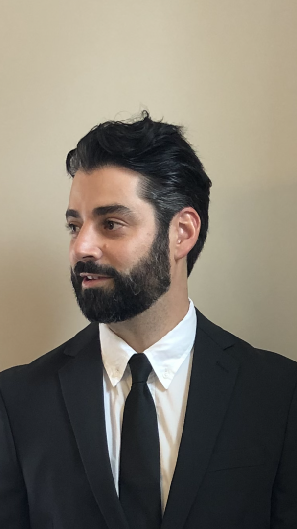 Man with styled dark hair and well-groomed full beard wearing a black suit, white dress shirt, and black tie
