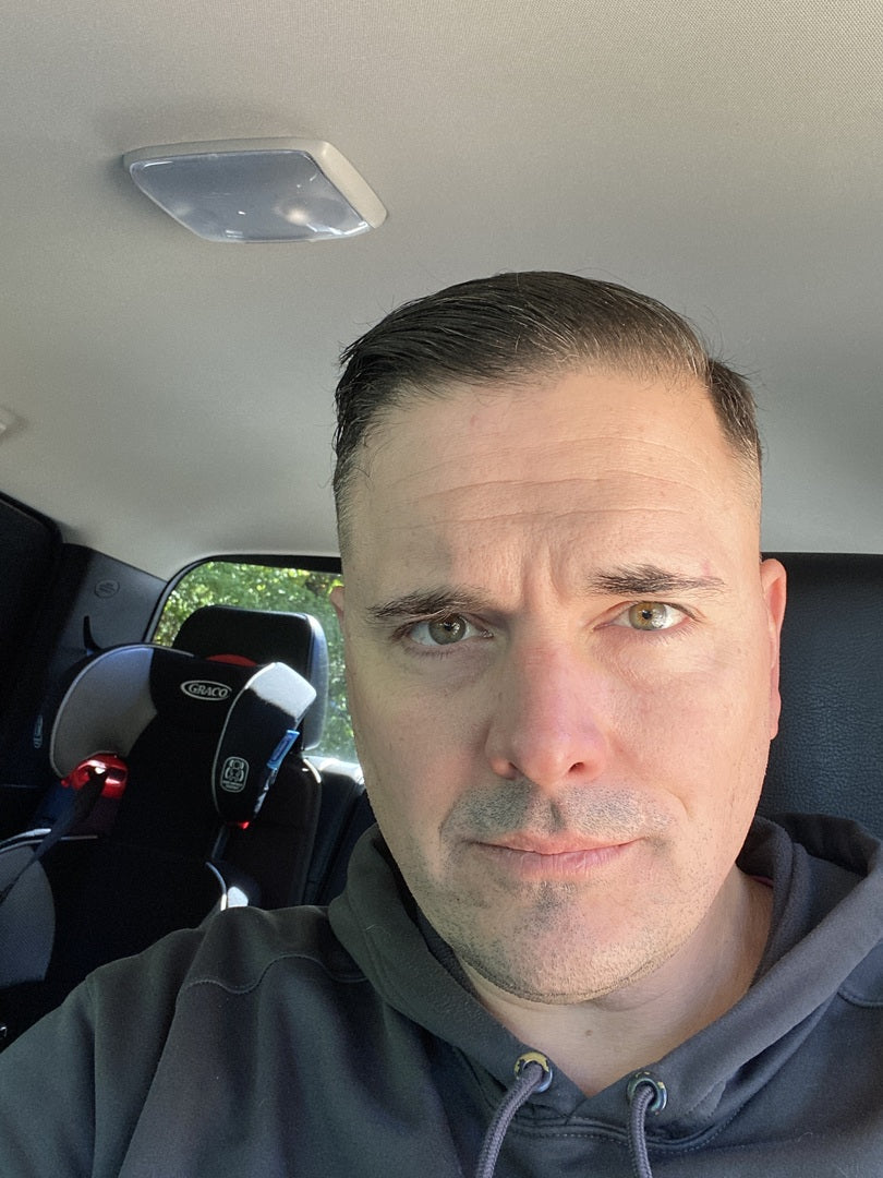 Man with short dark hair and light stubble in a car showing a fresh haircut, wearing a gray hoodie, Graco child seat behind him