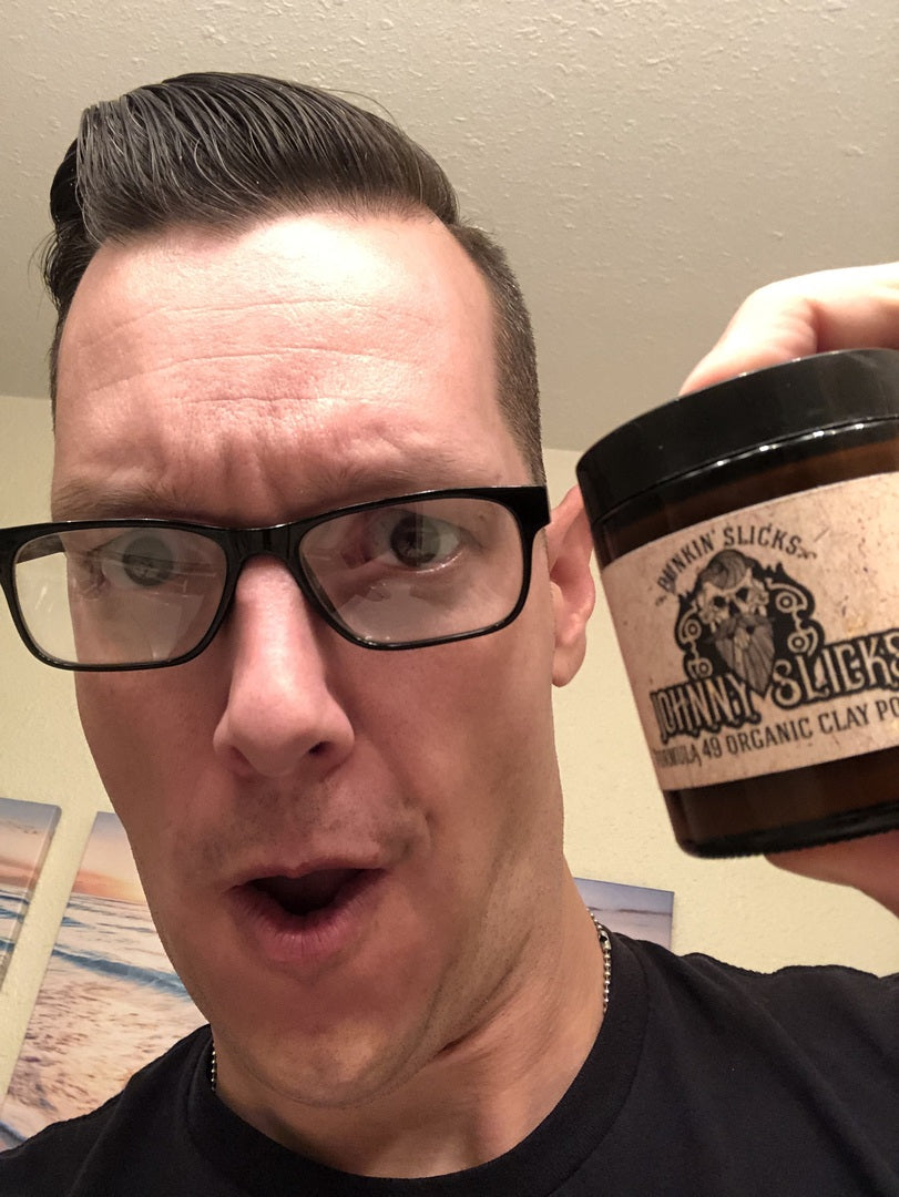 Man with slicked-back hair and glasses holding a jar of Johnny Slicks Formula 49 Organic Clay Pomade in amber container