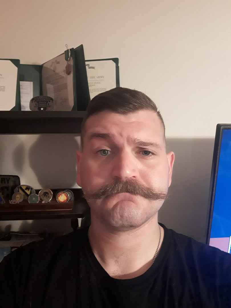 Man with thick styled mustache and short hair wearing black shirt, military challenge coins and Army certificate on shelf