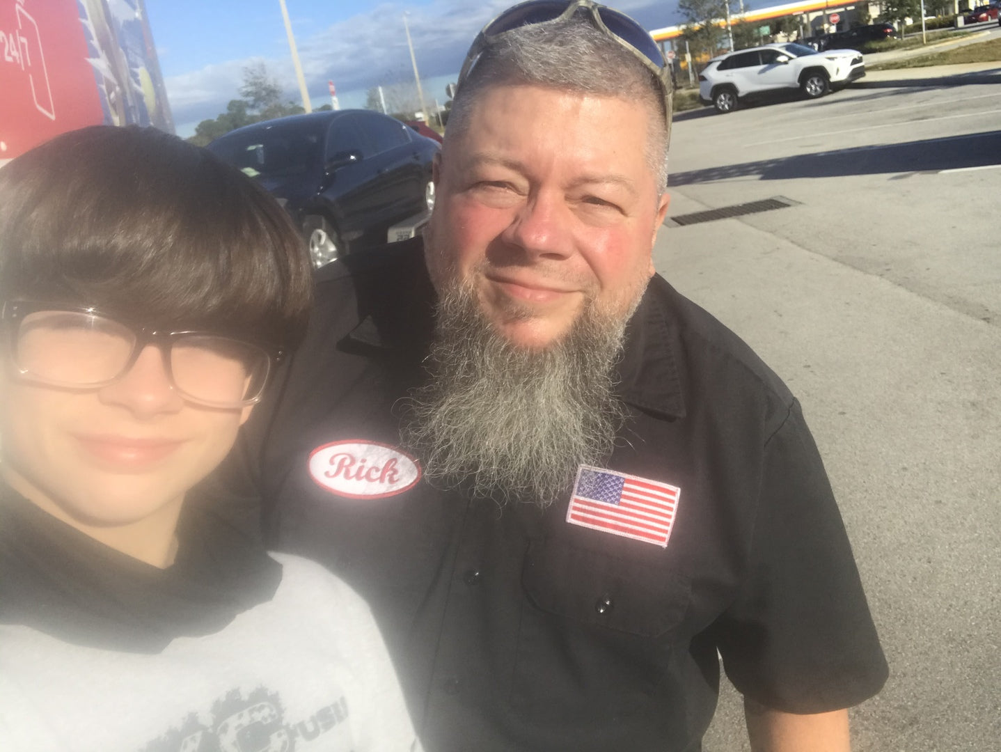 Man with long gray beard wearing black work shirt with "Rick" name patch and American flag patch, standing with young person outdoors