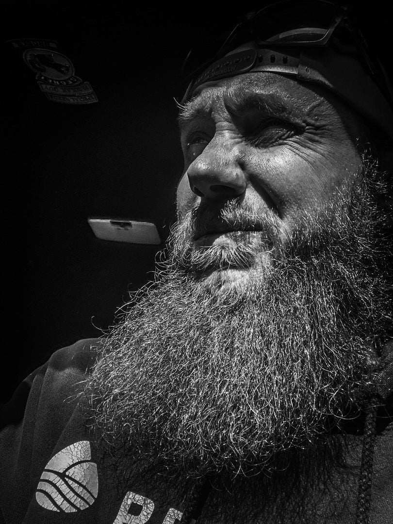 Man with long, full gray beard looking upward, wearing a cap and dark shirt with circular logo