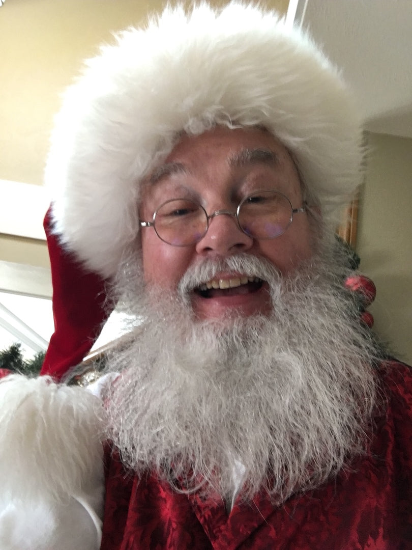 Man in Santa Claus costume with long full white beard, round glasses, red hat, and white gloves, smiling at camera