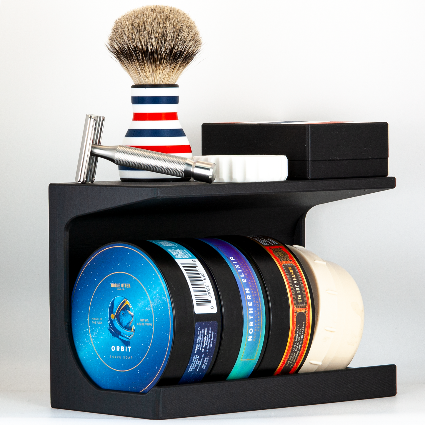 Black soap stand with a variety of Noble Otter shaving soaps on a white background. a razor, brush, storage case and razor refueler are stored on top.