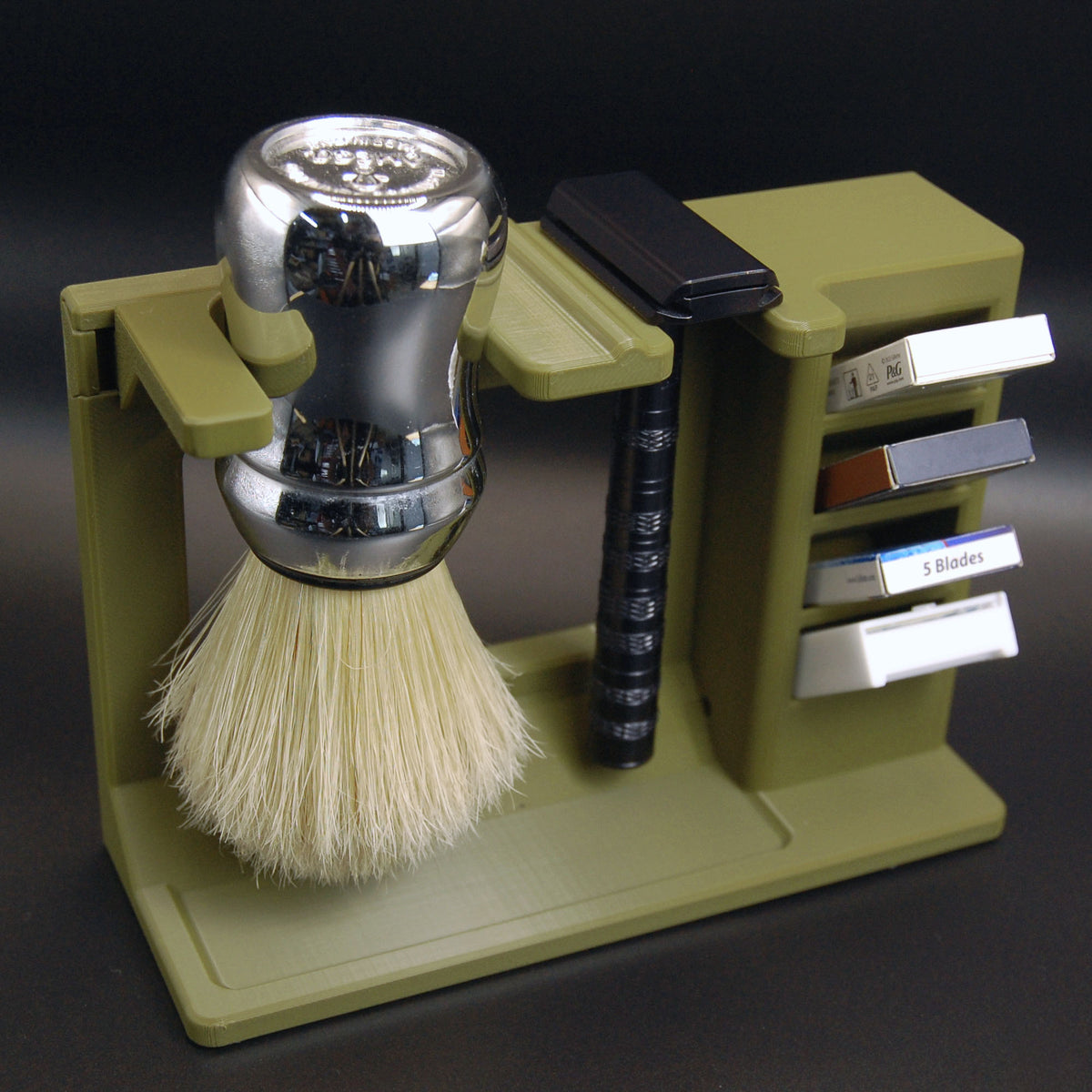 Army green 3D-printed stand with chrome Omega shaving brush, black adjustable safety razor, and four blade pack slots