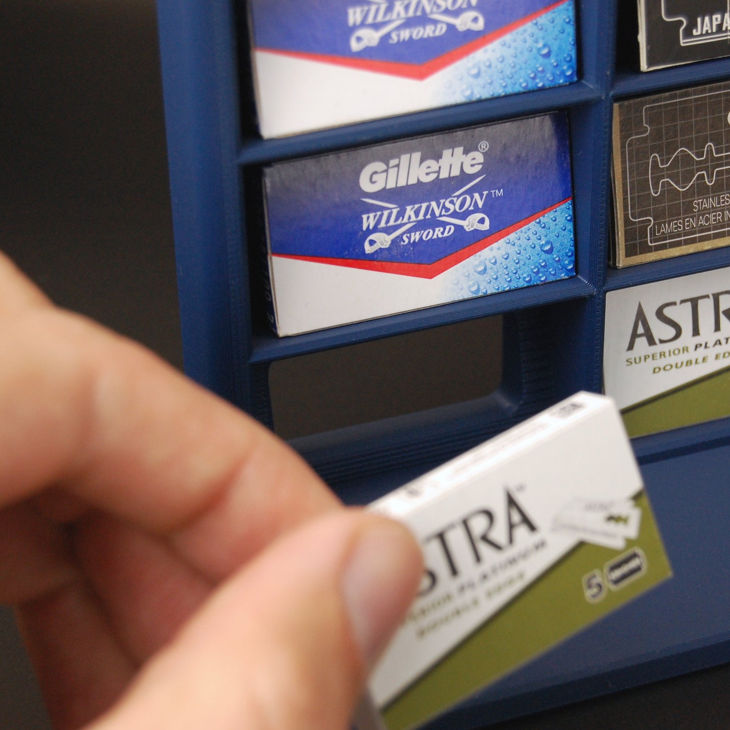 Various razor blade tucks stored in a navy blue razor blade display stand on a black background. Someone is grabbing a blade tuck showcasing a pocket for easy blade removal.