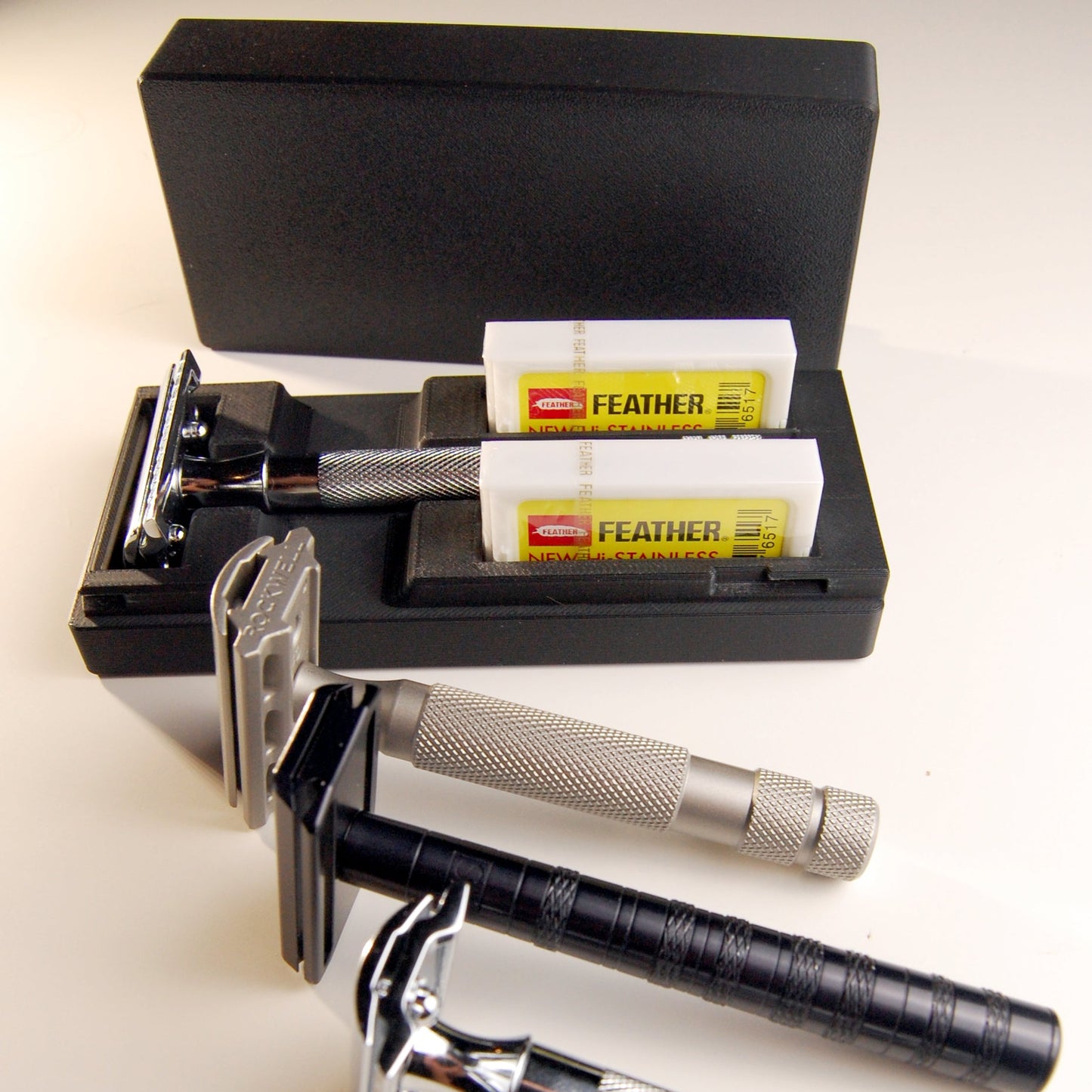 Black Universal razor case pictured with a Merkur 23C inside.  Three other razors are in front of the case.  A stainless steel Rockwell 6S, a black Henson AL13 and a chrome Merkur 34C.  
