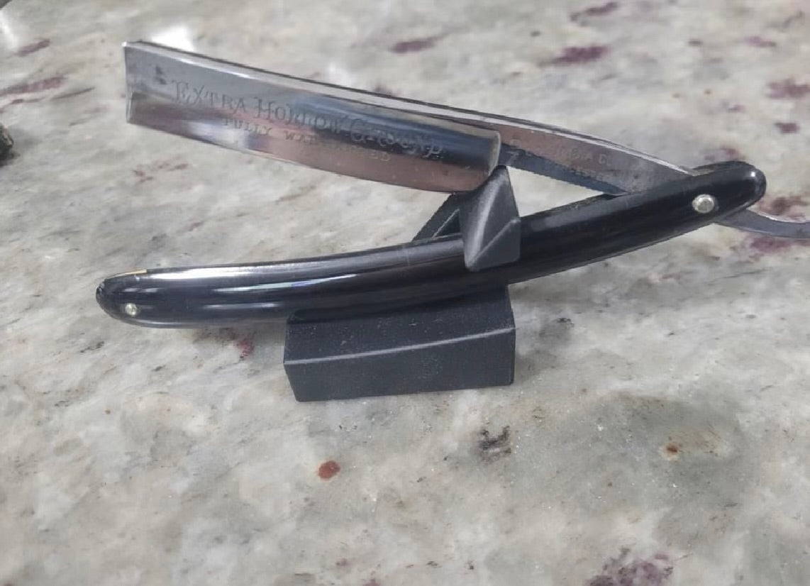 Black-handled straight razor with extra hollow ground blade resting open on The Launch Pad stand on a marble countertop