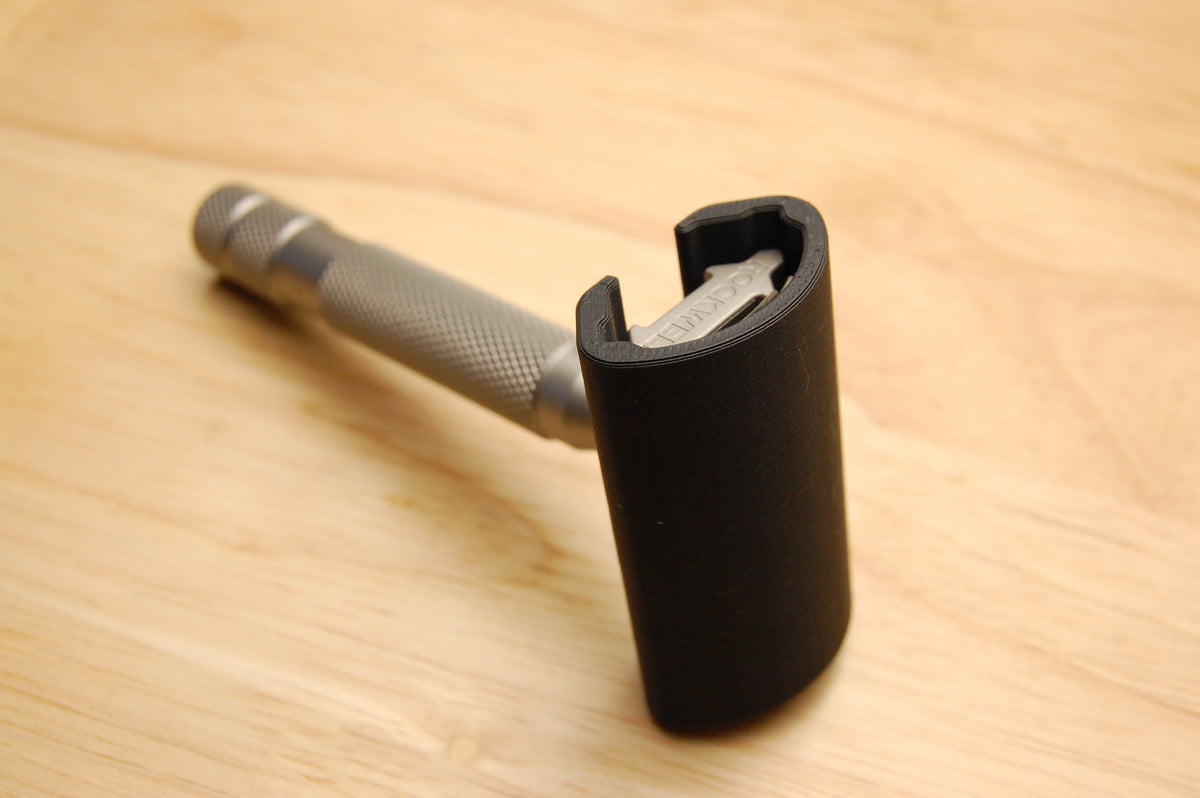 Black travel cover fitted over Rockwell safety razor head on wooden surface, showing snug protective fit over blade area