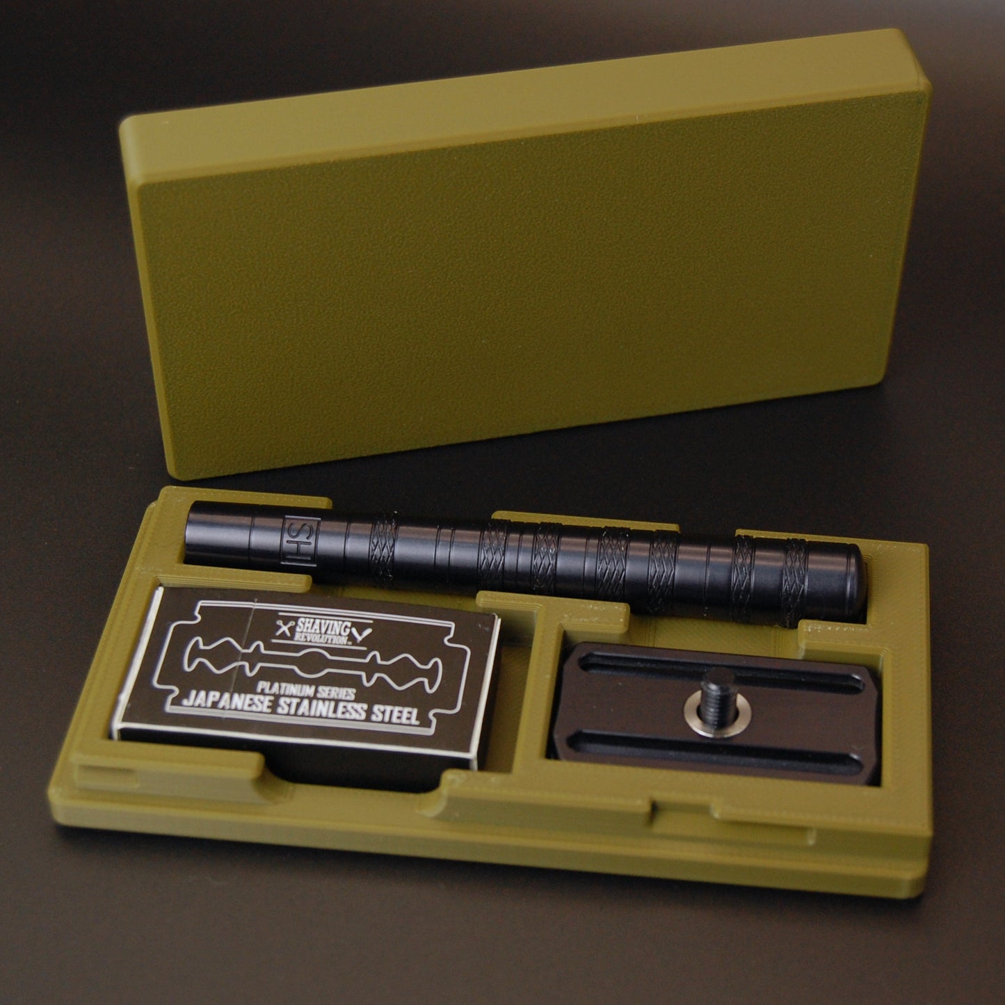 Henson case in army green on a black background.  Black Henson razor is stored inside with a blade tuck of Shaving Revolution Japanese stainless steel razor blades.