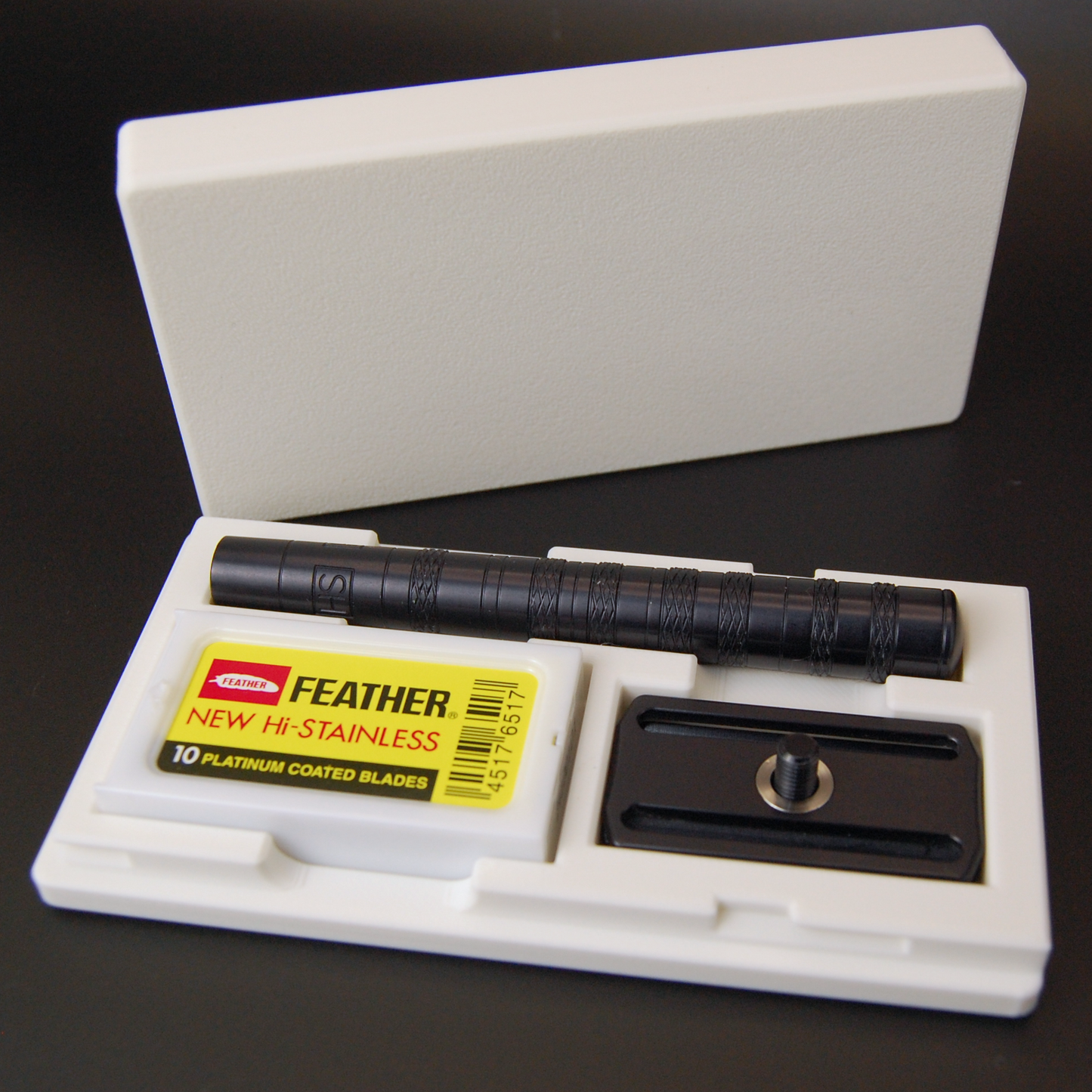 Henson case in white on a black background.  Black Henson razor is stored inside with a blade tuck of Feather New Hi-Stainless razor blades.