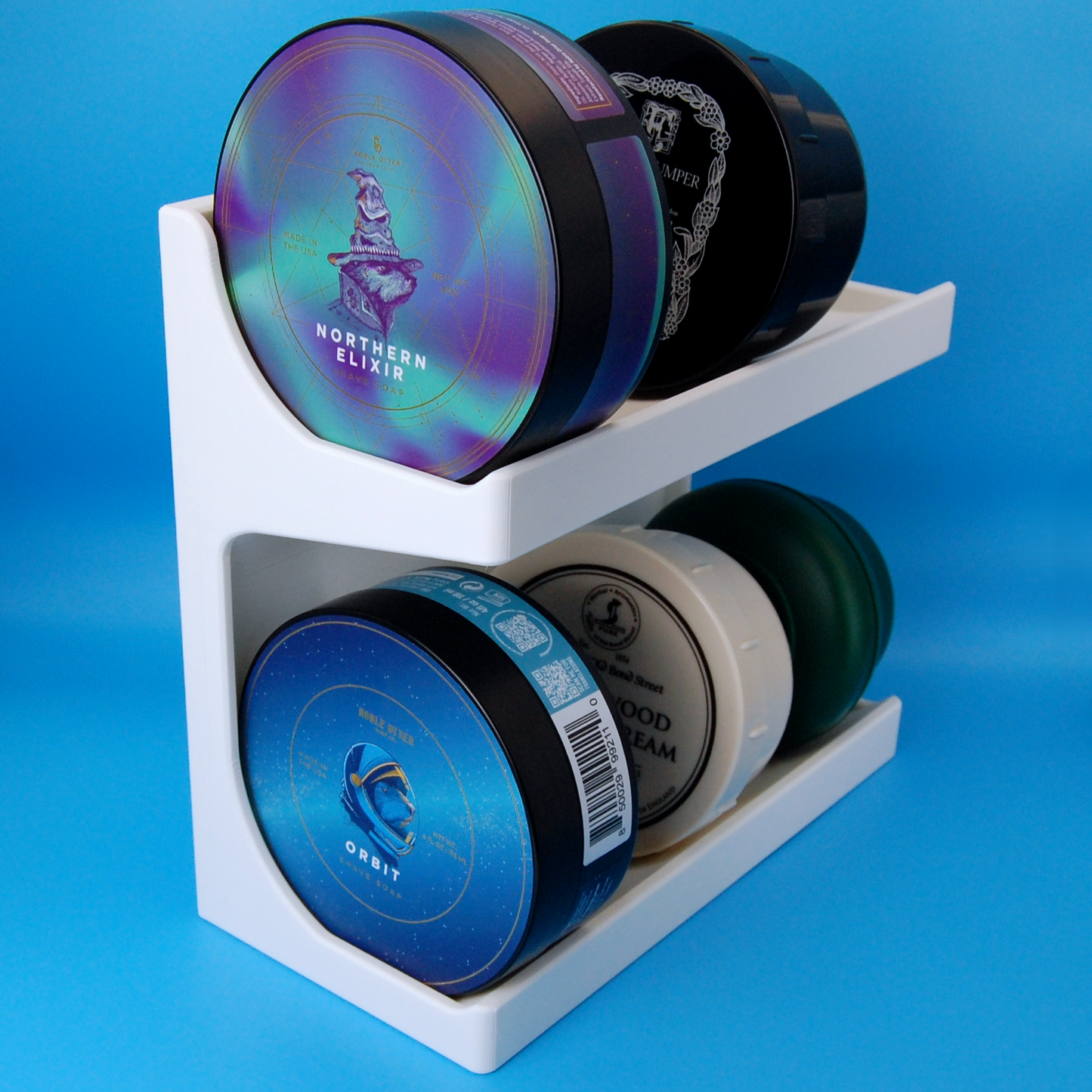 White shaving soap stand with several shaving soaps stored on it including two Noble Otter soaps, Orbit and Northern Elixir.  The stand is on a light blue background.
