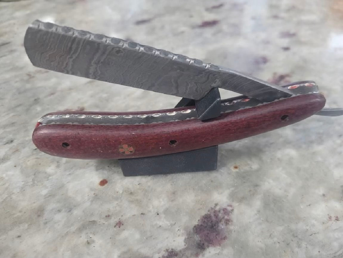 Straight razor with Damascus steel blade and dark red wood scales resting open on The Launch Pad stand on a marble countertop